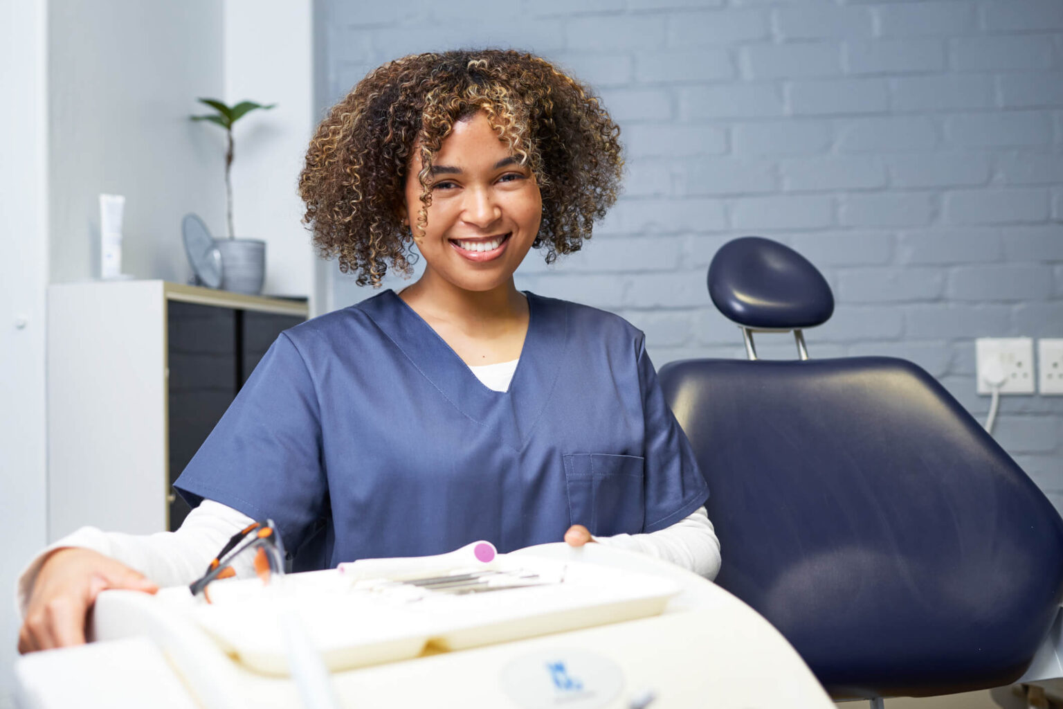 Expanded functions dental assistant in navy scrubs smiling beside a dental chair and instrument tray in a clinical office setting